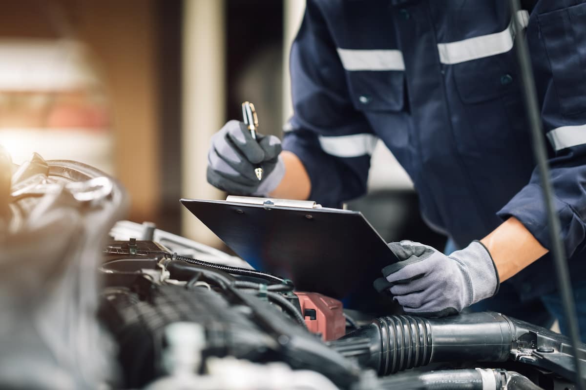 Mechanic wearing gloves and inspecting a car engine while writing notes on a clipboard.